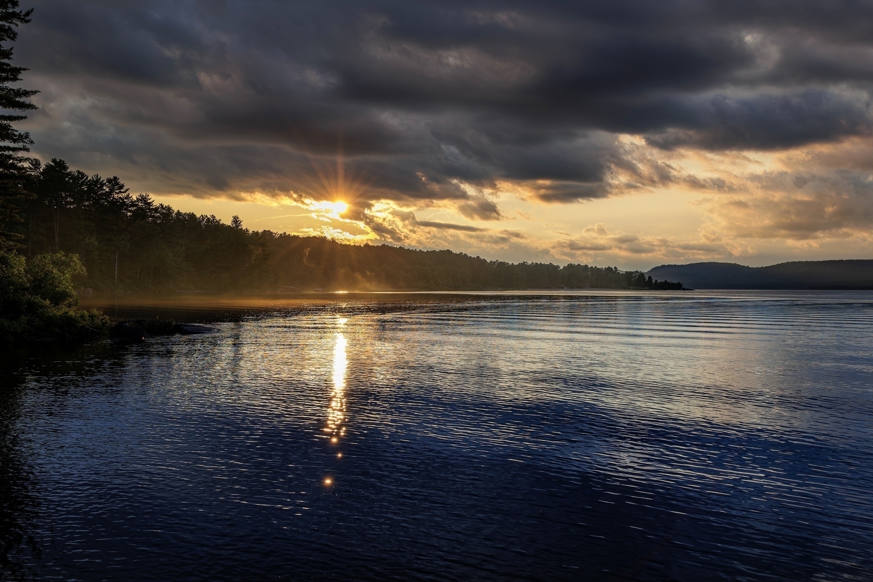 A serene river scene is captured with the sun setting behind clouds, casting reflections on the water.