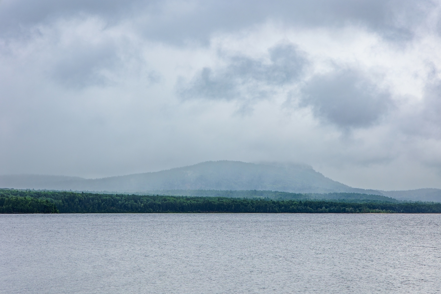 A calm river is framed by a distant tree line and a misty mountain under a cloudy sky.