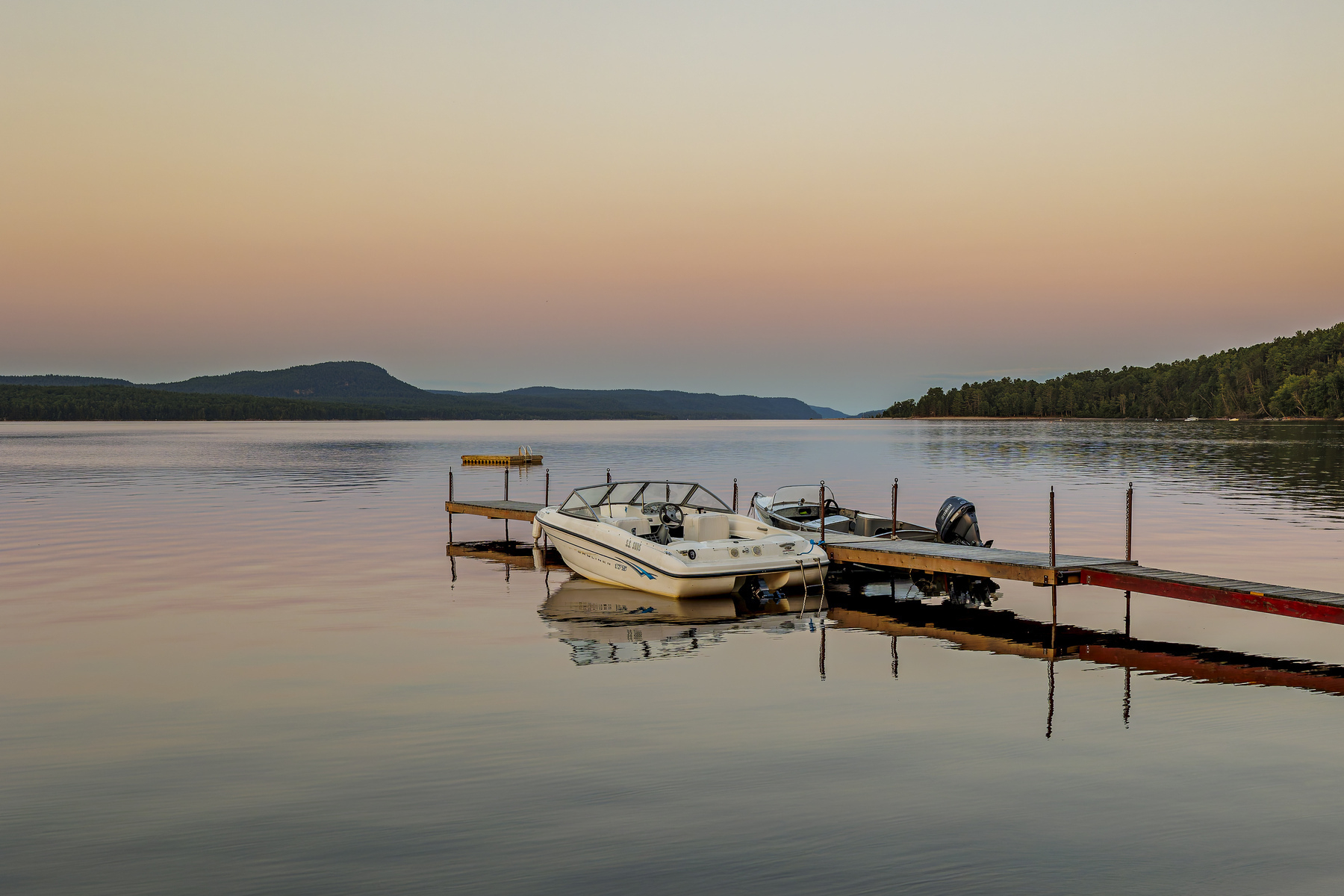 A small motorboat is docked on a peaceful river during a serene sunset, with distant hills and calm water reflecting the sky's colors.