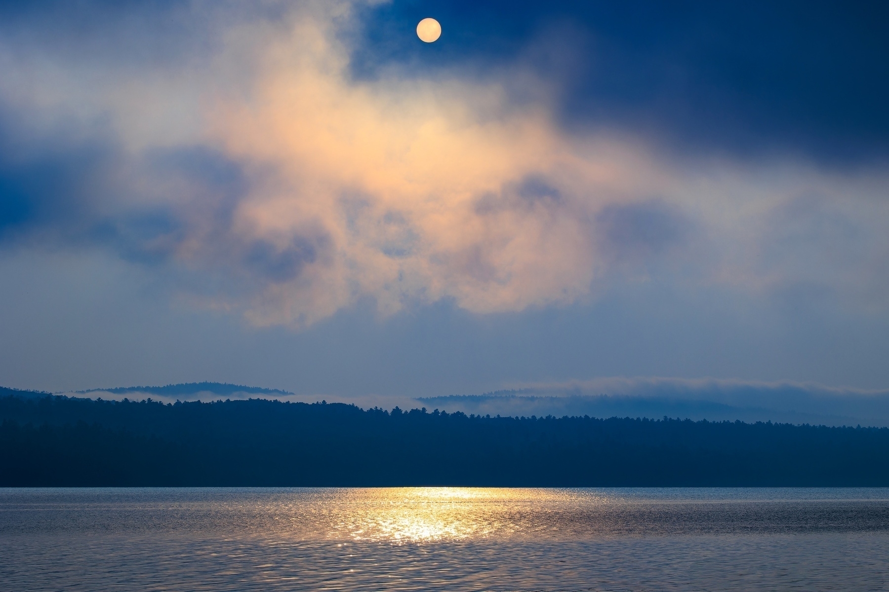 A serene river reflects the soft glow of the rising sun through misty clouds and over distant hills.