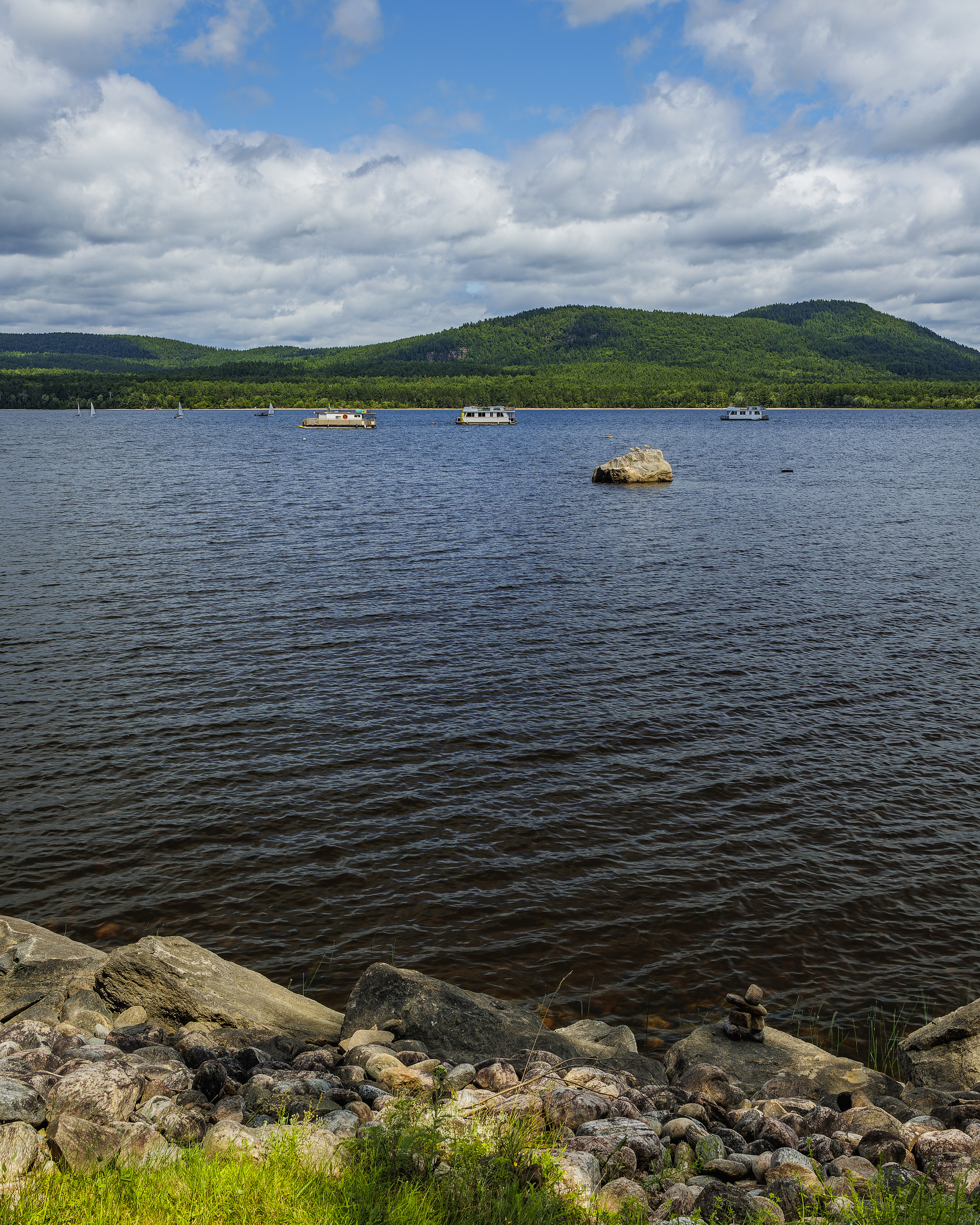 A serene river with a rocky shoreline is framed by distant, tree-covered hills under a partly cloudy sky.