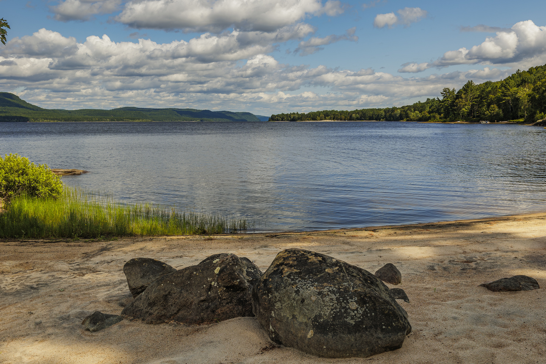 A serene riverside scene features clear blue water, a sandy shore with large rocks, and a backdrop of lush green trees under a partly cloudy sky.